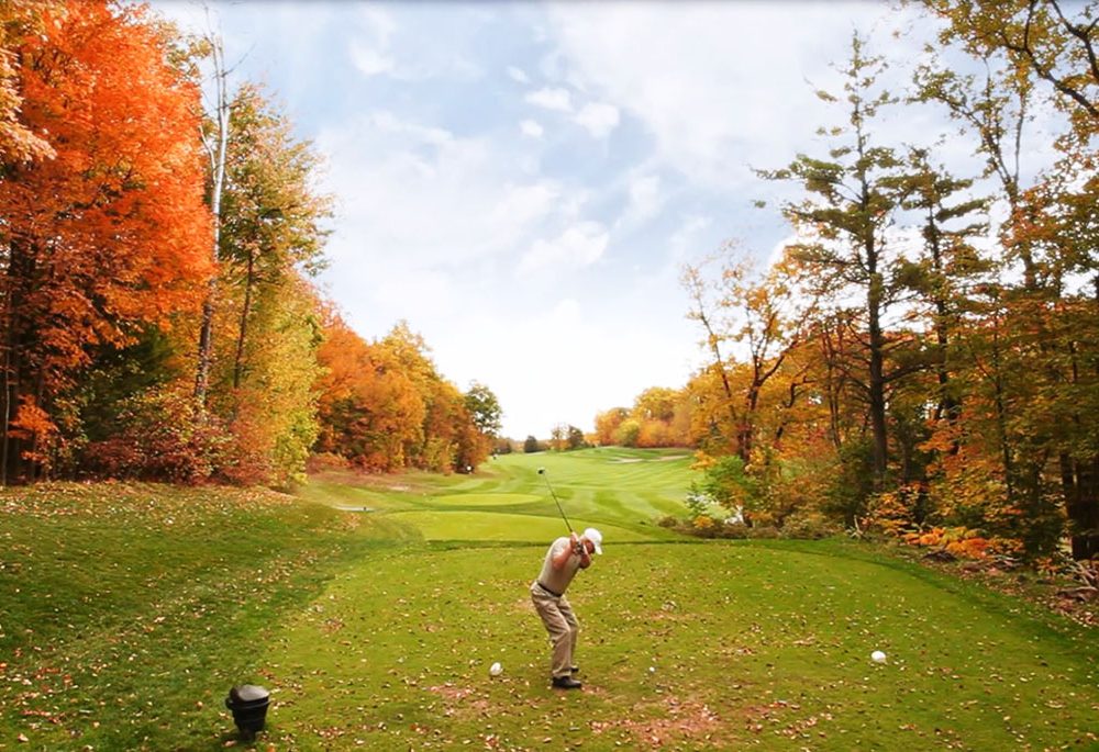 A person tees off at a golf course surrounded by orange leaved trees